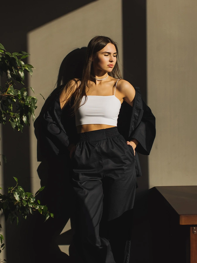 Woman in white crop top and black oversized coat leaning against wall in natural sunlight with plants - Breast reduction in Owings Mills, MD