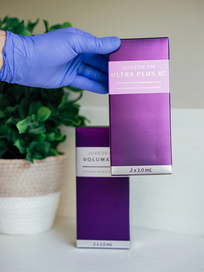 Hand in blue glove holding purple and white cosmetic product boxes next to potted plant on white surface - Juvederm in Owings Mills, MD