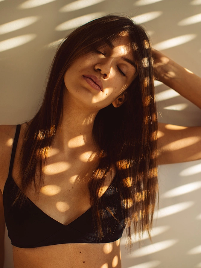 Woman with long dark hair in black top with sunlight shadows streaming through window blinds - Lateral chest skin removal in Owings Mills, MD