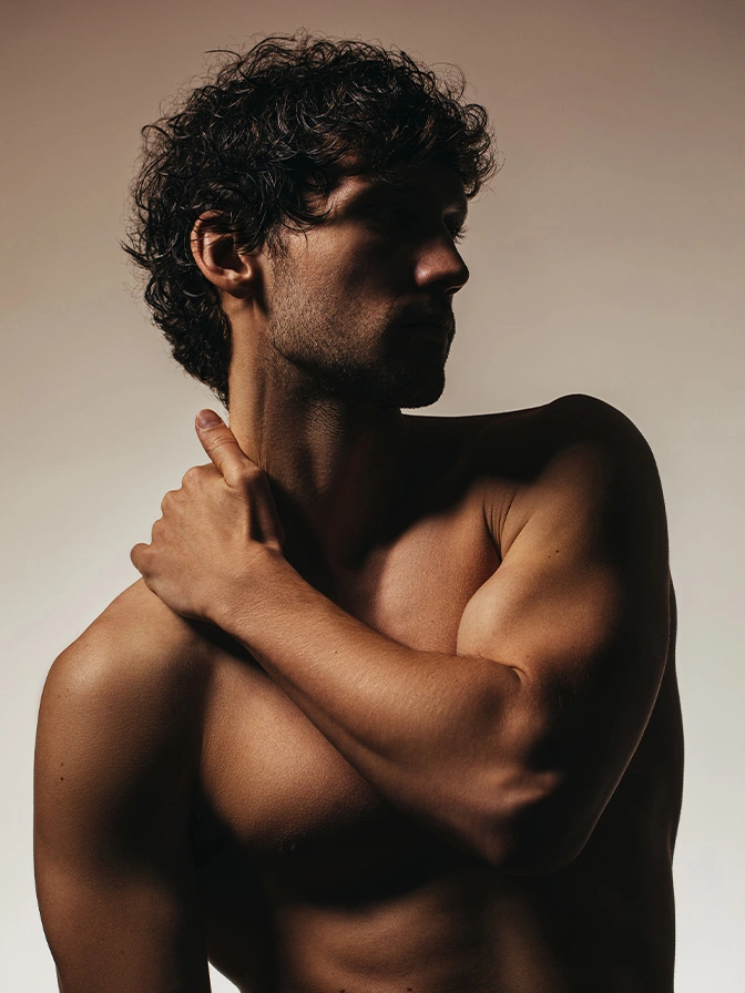 Shirtless man with curly hair touching shoulder in profile against neutral background - Liposuction in Owings Mills, MD